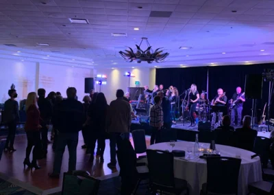 A lively band performs on stage as an audience dances and enjoys the music in a warmly lit venue with round tables.