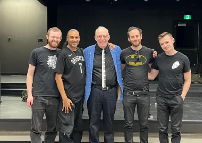 Five men pose together in a dimly lit theater, dressed in black attire, showcasing a mix of casual and themed outfits.