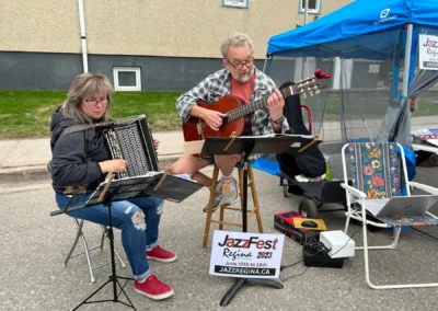 Musicians play at the Jazz-Fest Regina 2023, featuring an accordionist and a guitarist, with a sign promoting the event nearby.