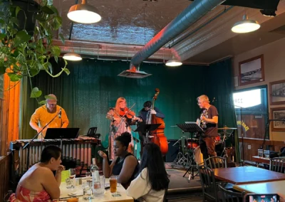 A lively indoor music performance featuring four musicians at a bar, with patrons enjoying drinks at tables in the foreground.