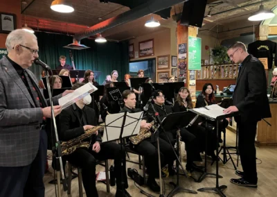 A conductor leads a band of musicians, seated with various instruments, in a lively pub setting illuminated by warm lights.